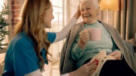 Caregiver gently pats an elderly woman’s head as they sit together; the senior holds a pink mug and wears a teal sweater with a blanket on her lap.