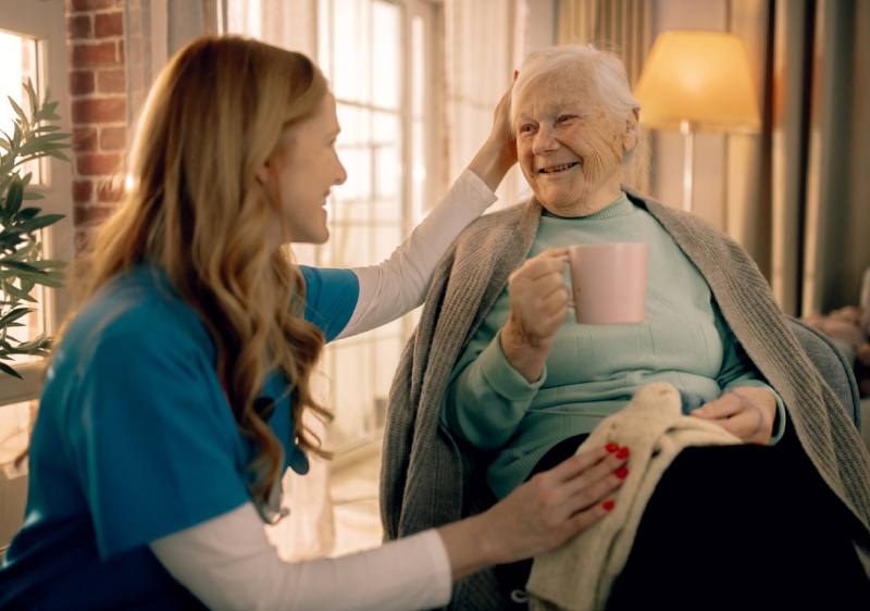 Caregiver gently pats an elderly woman’s head as they sit together; the senior holds a pink mug and wears a teal sweater with a blanket on her lap.